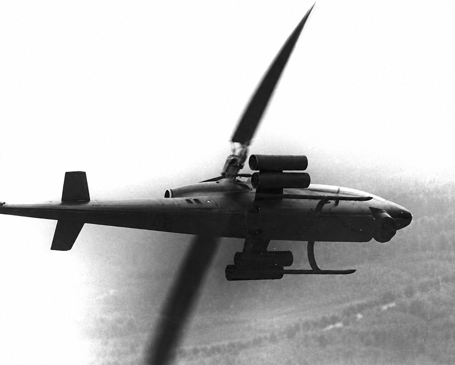 Black-and-white image of a Bell AH-1 Cobra attack helicopter banking in flight with rocket pods mounted on its stub wings during Vietnam War operations.