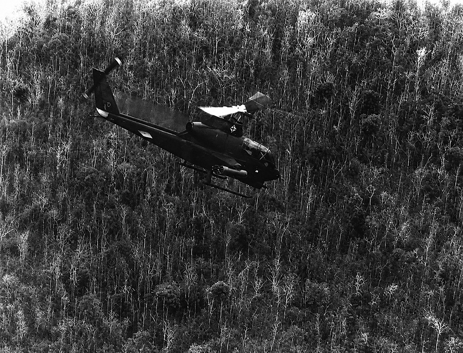 Black-and-white aerial image of a Bell AH-1 Cobra attack helicopter flying low above dense jungle terrain during Vietnam War combat operations.