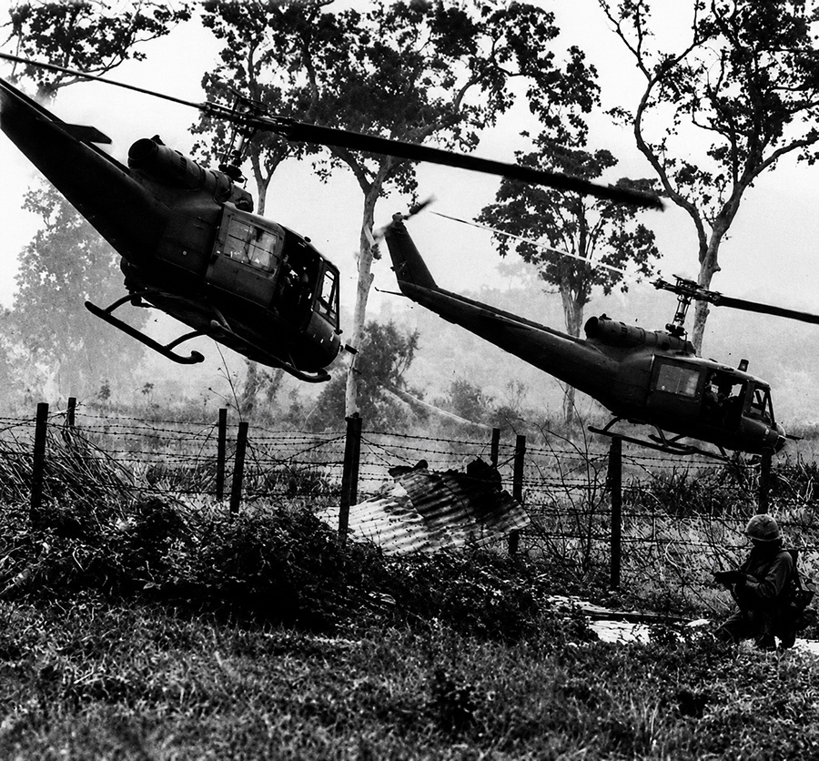 Two Bell UH-1 Huey helicopters flying low over a Vietnam War landing zone with troops positioned near jungle terrain and barbed wire.