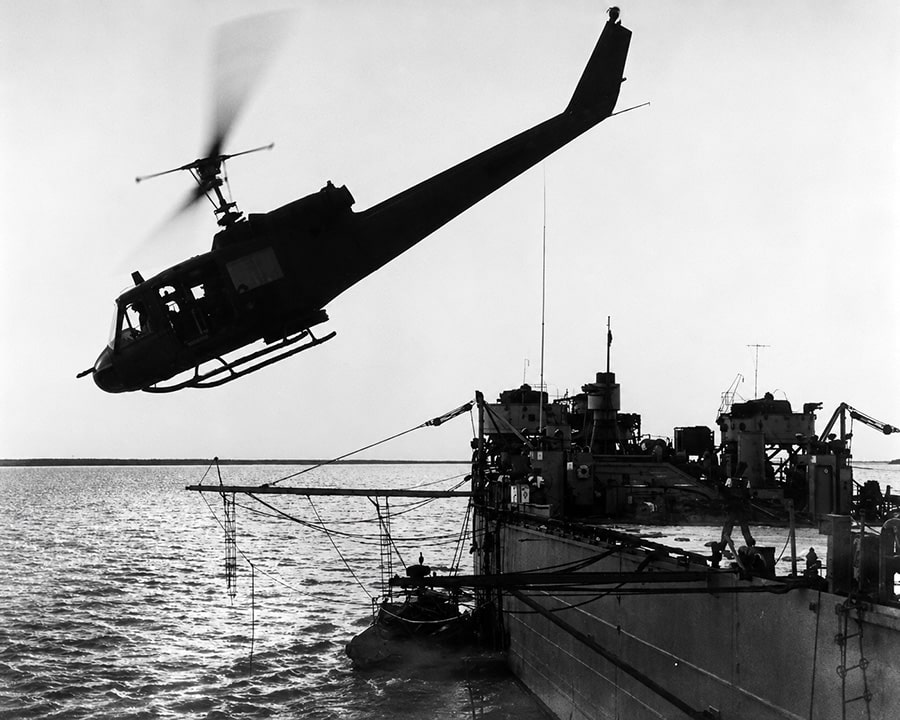 Black-and-white image of a Bell UH-1 Iroquois “Huey” helicopter hovering above a military supply ship during Vietnam War operations, highlighting the aircraft’s troop transport and air assault role.
