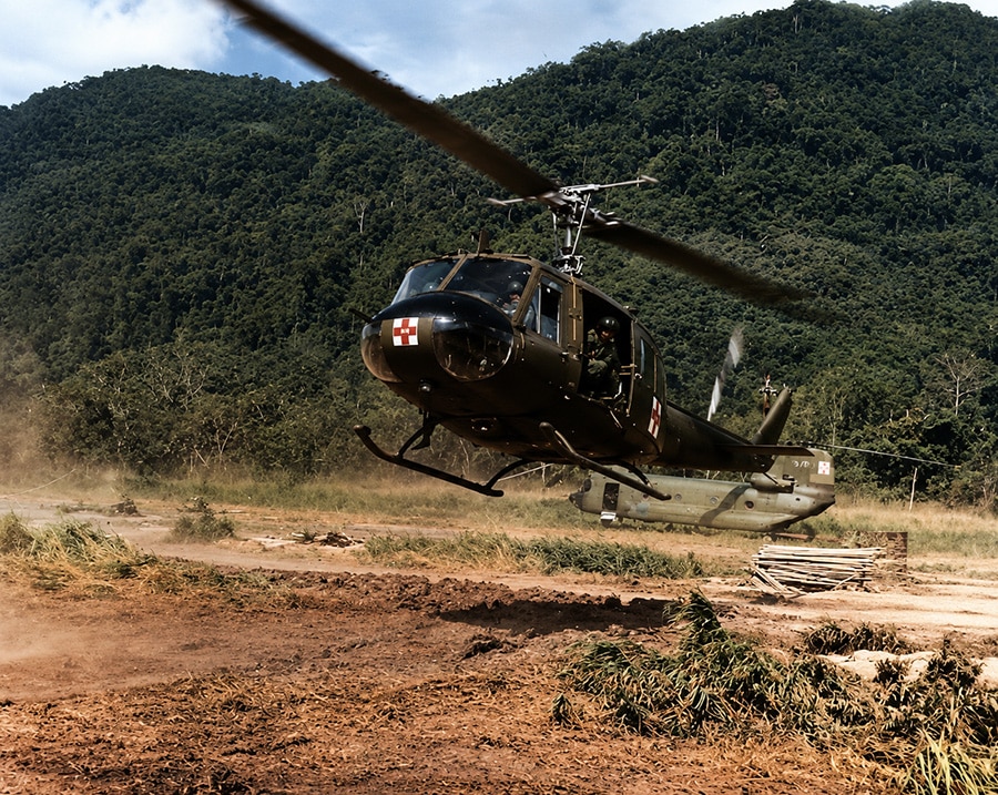 Bell UH-1 Huey Dustoff medevac helicopter landing in a dusty Vietnam clearing with jungle-covered mountains behind it.
