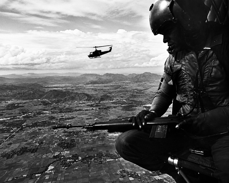 Black-and-white image of a Huey door gunner watching the landscape from an open helicopter during a Vietnam War flight.