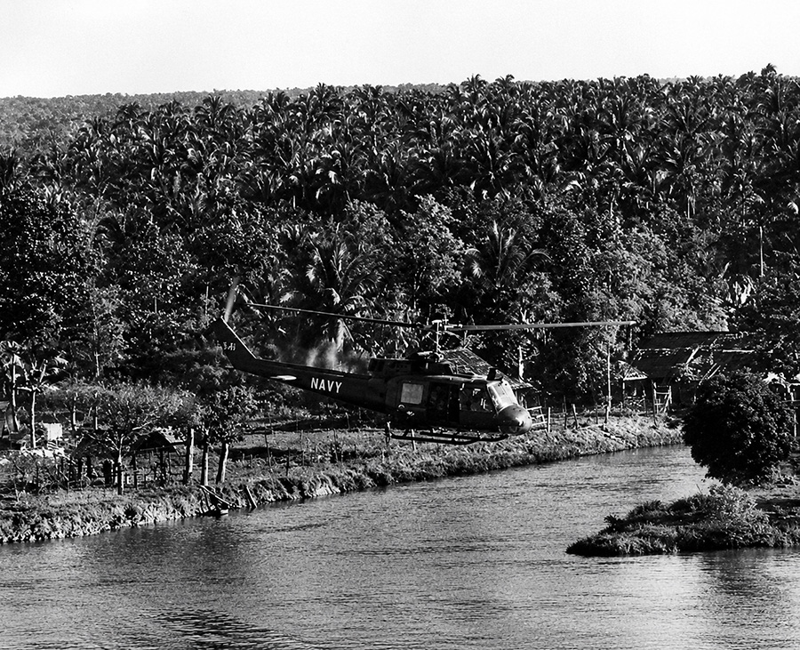 Black-and-white image of a U.S. Navy Bell UH-1 Huey gunship flying low over a canal beside a village and dense vegetation in Vietnam.