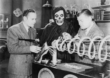 The Crimson Ghost stands beside the Cyclotrode ray gun with two men in a scene from the 1946 Republic serial